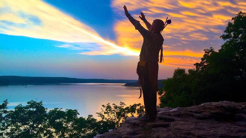 man reaching up to the sky in front of a sunset