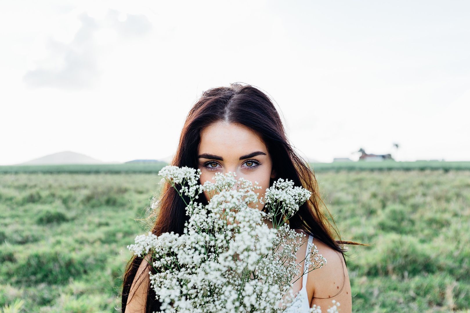 Junge aussehende Frau steht vor einem Feld und verdeckt ihr Gesicht zur Hälfte mit Blumen, sie hat braune Augen und lange braune Haare